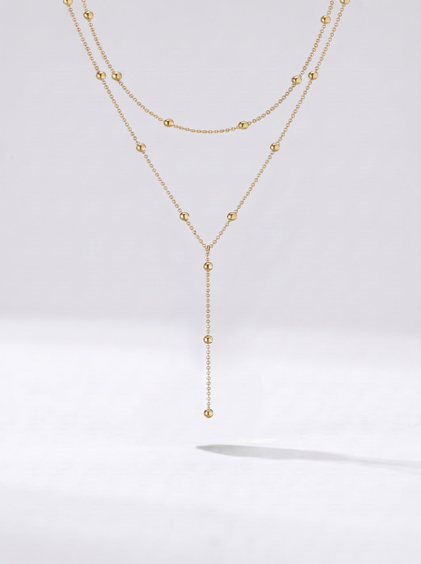 Colar dourado comprido em camadas com detalhes em esferas e pendente delicado, fotografado sobre fundo claro para um visual elegante, sofisticado e moderno.