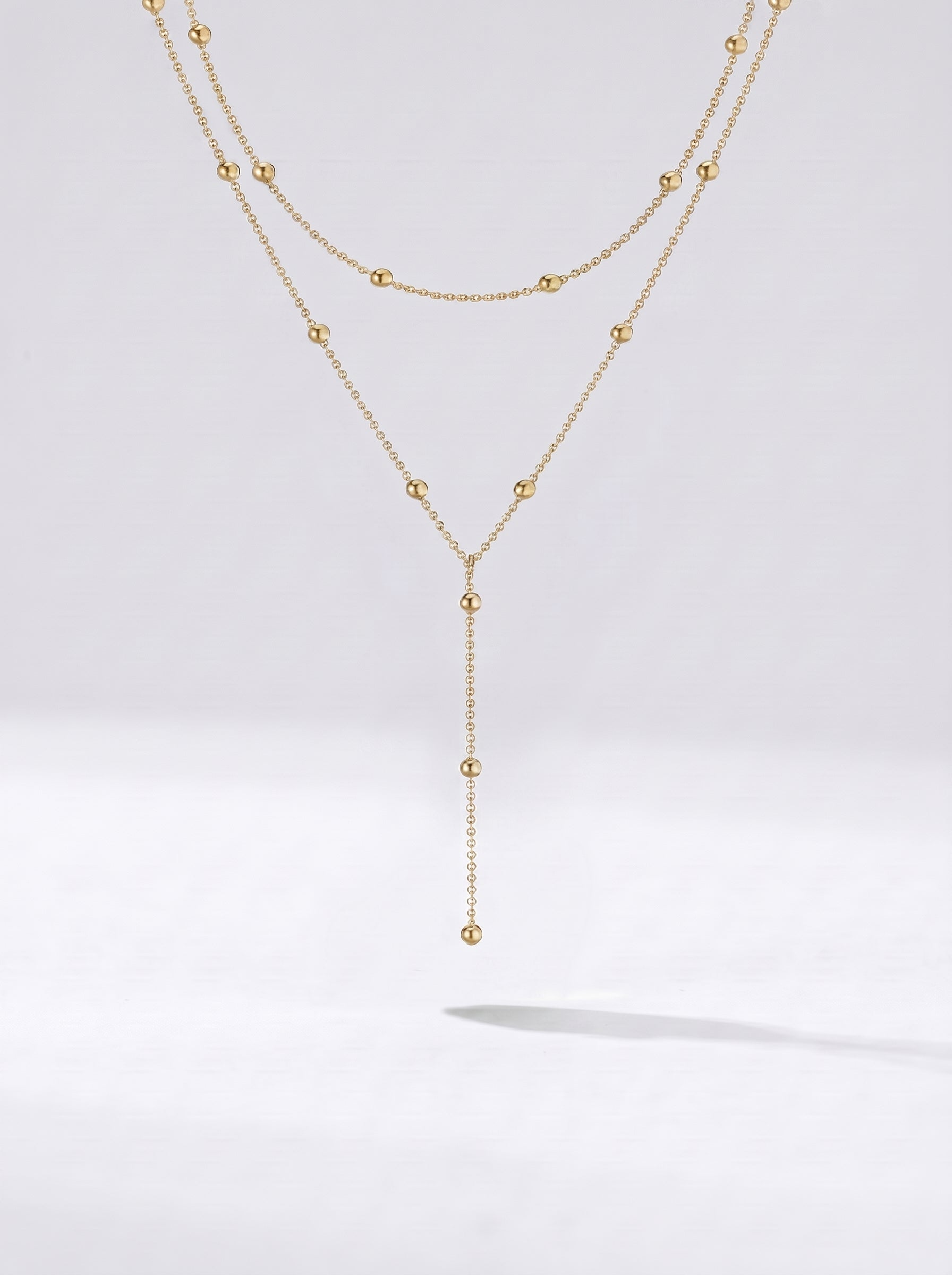 Colar dourado comprido em camadas com detalhes em esferas e pendente delicado, fotografado sobre fundo claro para um visual elegante, sofisticado e moderno.