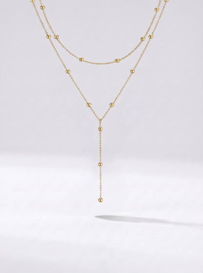 Colar dourado comprido em camadas com detalhes em esferas e pendente delicado, fotografado sobre fundo claro para um visual elegante, sofisticado e moderno.