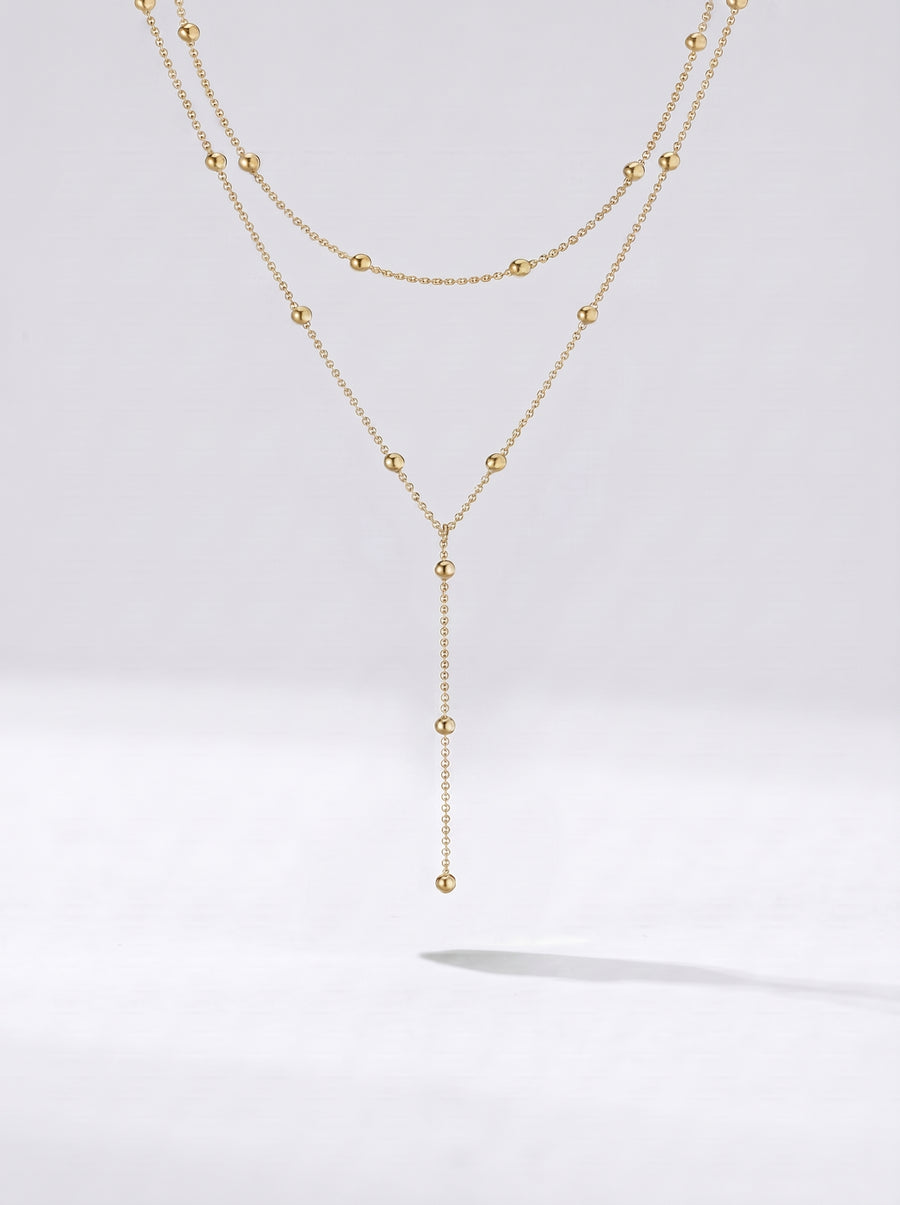 Colar dourado comprido em camadas com detalhes em esferas e pendente delicado, fotografado sobre fundo claro para um visual elegante, sofisticado e moderno.